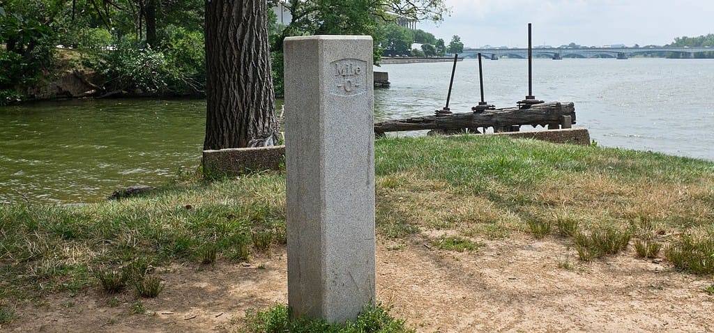 Stone post at the start of the Chesapeake and Ohio Canal in DC labeled Mile 0, with Kennedy Center and Roosevelt Bridge in the background. Credit: Bonnachoven, Wikimedia Commons