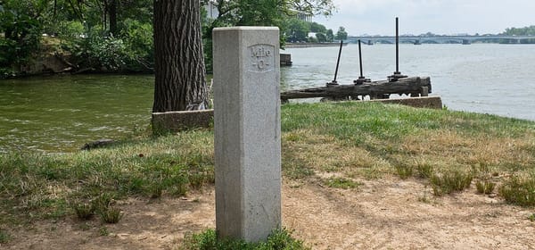 Stone post at the start of the Chesapeake and Ohio Canal in DC labeled Mile 0, with Kennedy Center and Roosevelt Bridge in the background. Credit: Bonnachoven, Wikimedia Commons