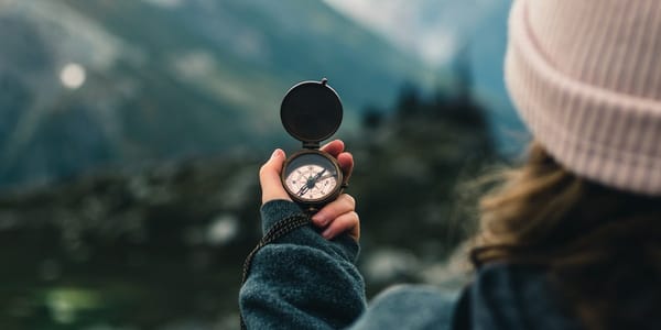 A white woman, viewed from behind her shoulder, looks out at an out-of-focus hiking landscape while holding a pocket compass.